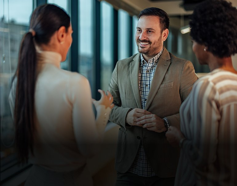 Smiling businessman in a beige blazer talking with two female colleagues during a meeting in a modern glass office.