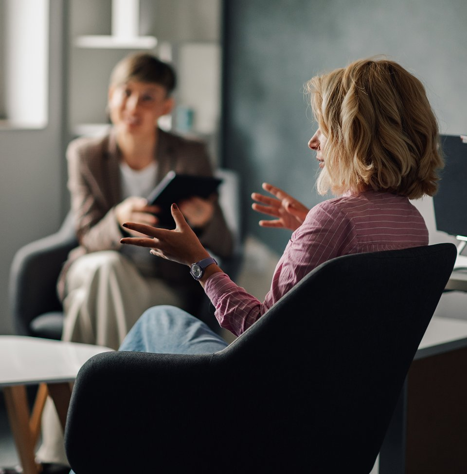 Woman in pink blouse gesturing while talking to a counselor with a tablet during a one-on-one therapy or coaching session.