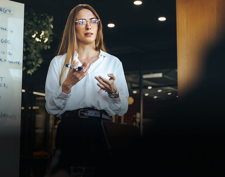 Professional woman in white blouse presenting business strategy notes from a whiteboard during a team meeting.