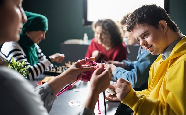 Group of young adults including a man with Down syndrome doing crafts and knitting together at a community workshop.